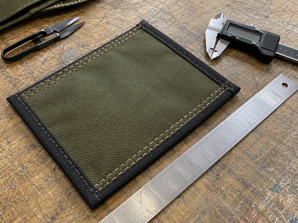 Seam and dimension checks | Fort Yuma Defense Co. Bound olive fabric sample with parallel stitching beside a steel ruler and digital caliper on a workbench.